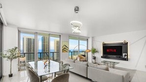 Een moderne woonkamer met een glazen eettafel, witte banken, een aan de muur bevestigde televisie en een balkon met uitzicht over de stad.