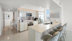 Een moderne keuken met witte kasten, een centraal eiland met barstoelen, roestvrijstalen apparatuur en een groot raam dat voor natuurlijk licht zorgt.