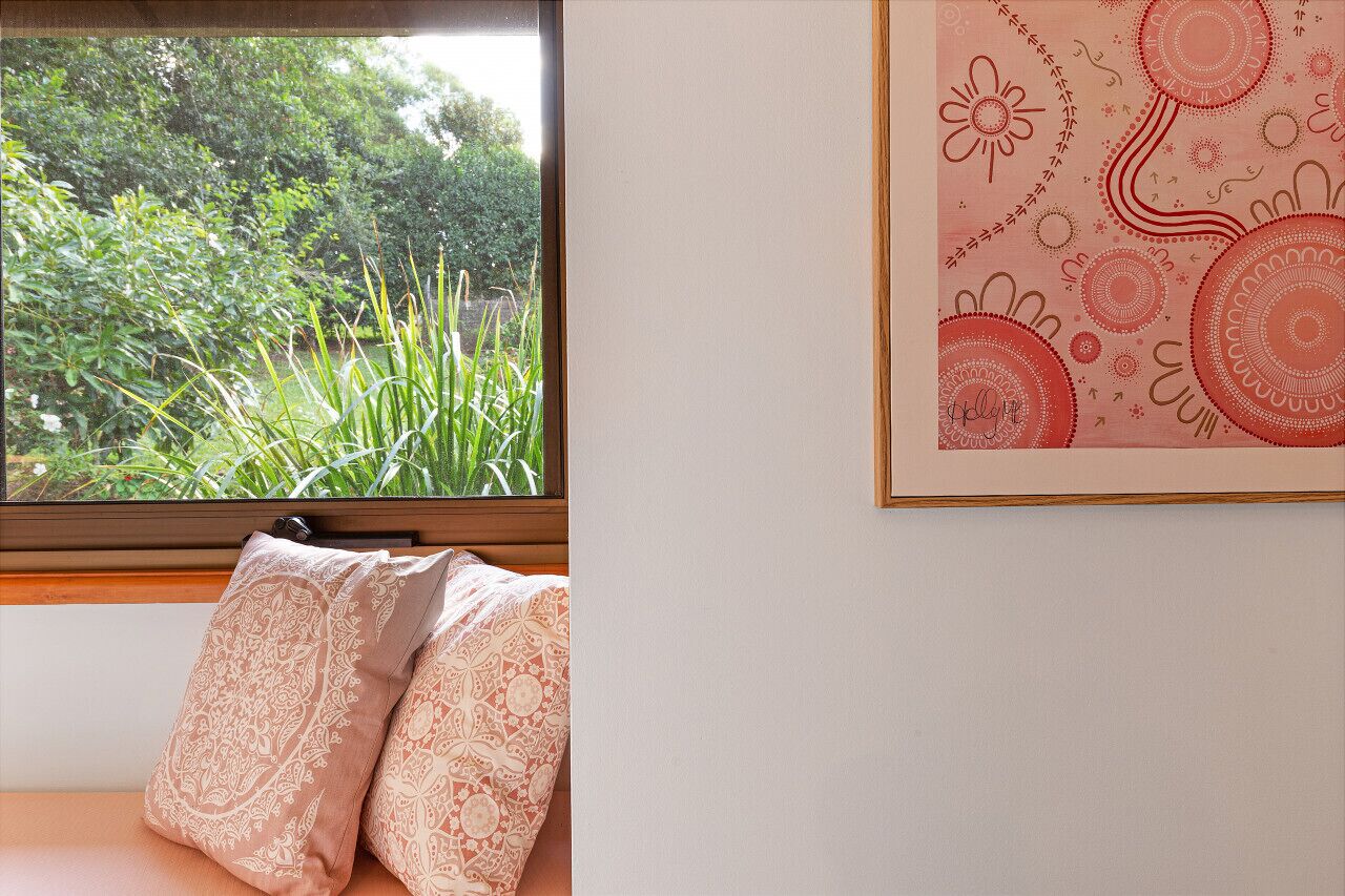 Een kamer met een raam dat uitkijkt op groen, een bed met gekleurde kussens en een muur met een lijstwerk abstract kunstwerk.