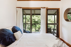 Relax in a serene bedroom with a large bed, wooden ceiling beam, and a view of lush greenery outside.