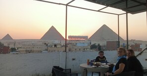 A rooftop terrace with a view of the pyramids, a table with food and drinks, and a metal structure in the background.