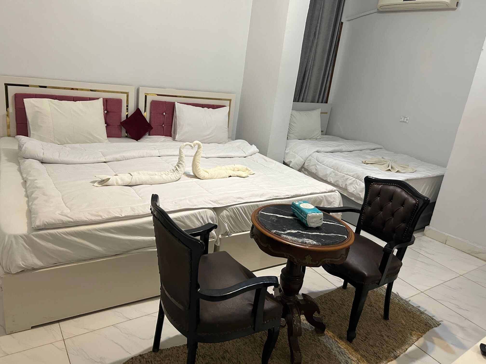 Une chambre d’hôtel avec un grand lit, deux petits lits, une table ronde avec des chaises et une petite table avec une bouteille.