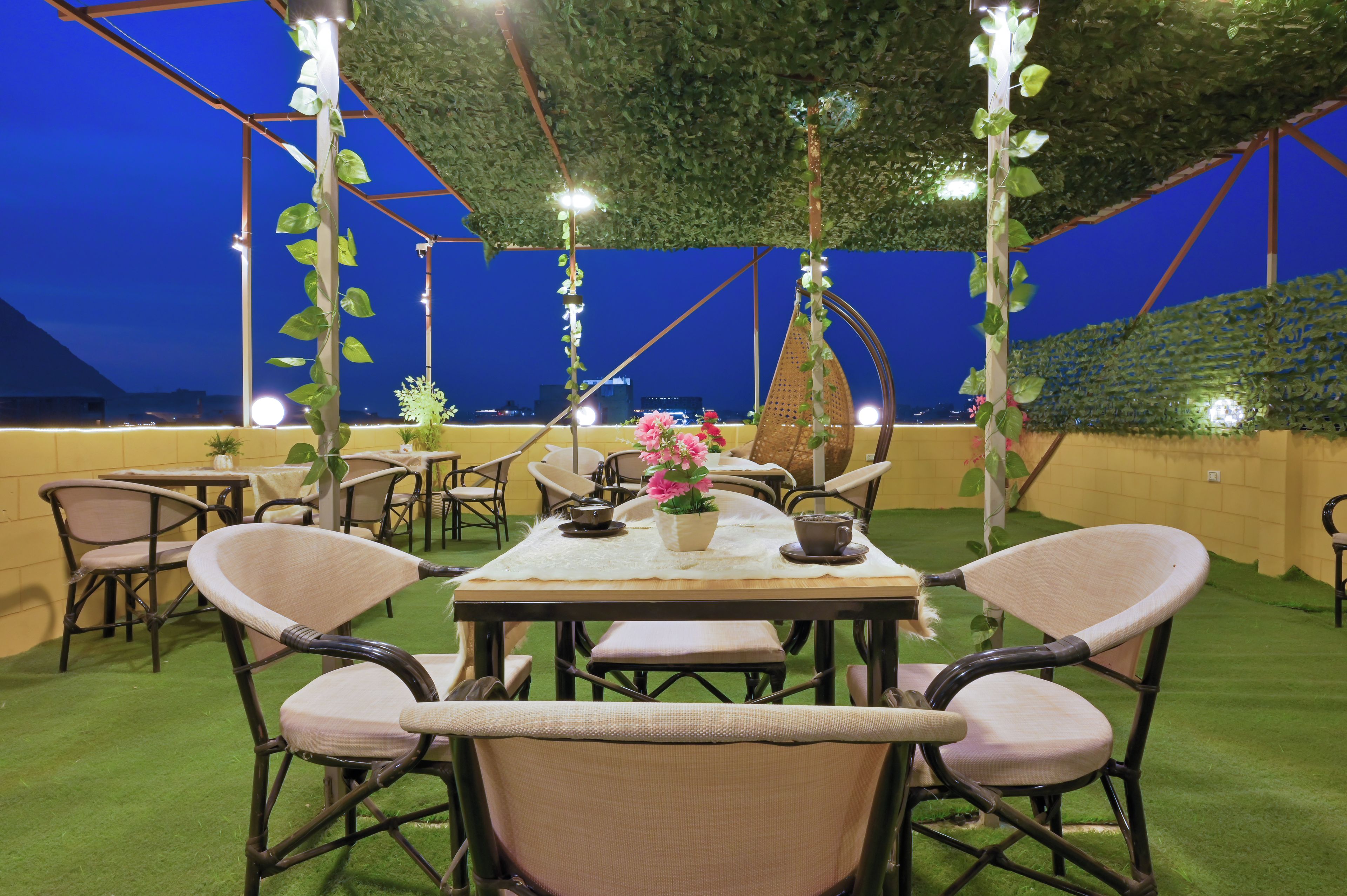 An outdoor dining area with tables and chairs, surrounded by greenery and a clear sky.