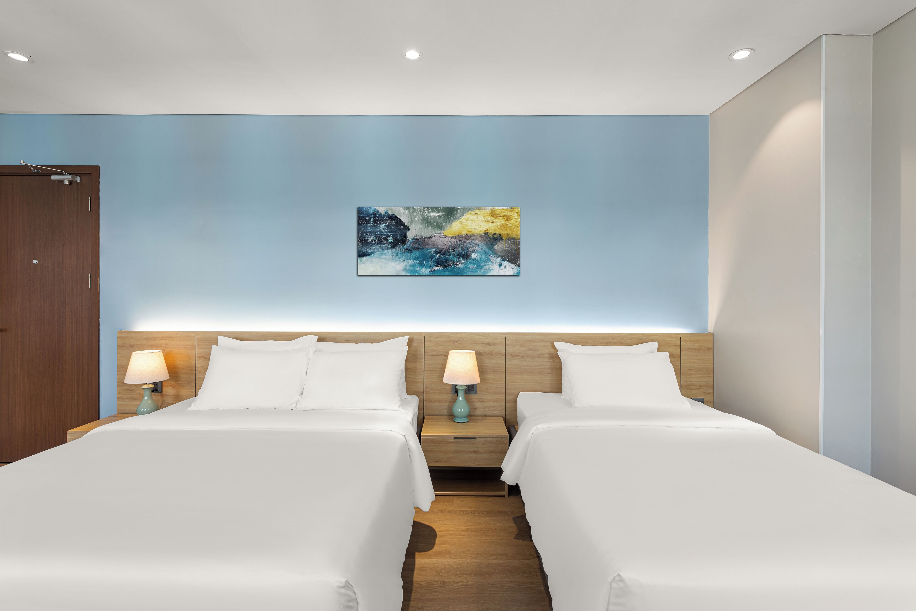 A hotel room with two beds, a wooden headboard, a painting on the wall, and a nightstand with a lamp.