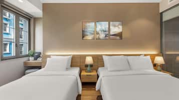 A hotel room with two beds, a wooden headboard, a nightstand with a lamp, and a window with a view of buildings.