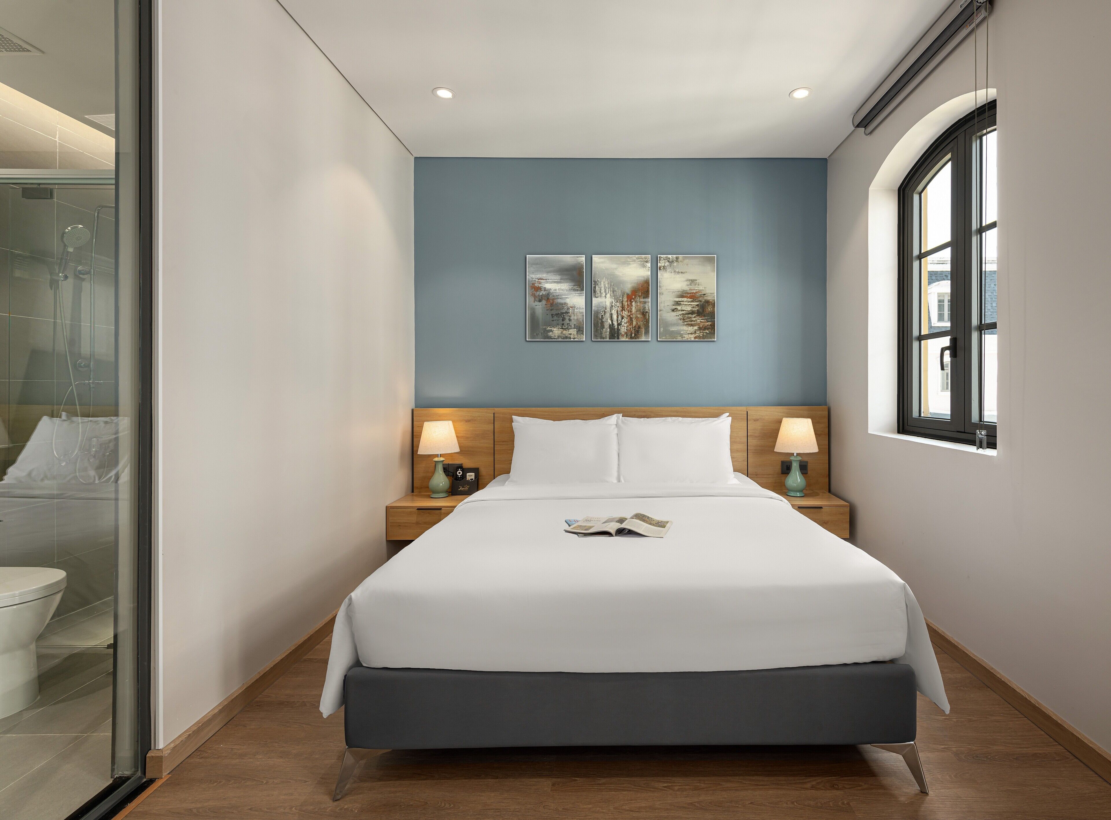 Habitación de hotel con cama, dos mesitas de noche con lámparas, una ventana y una obra de arte en la pared.