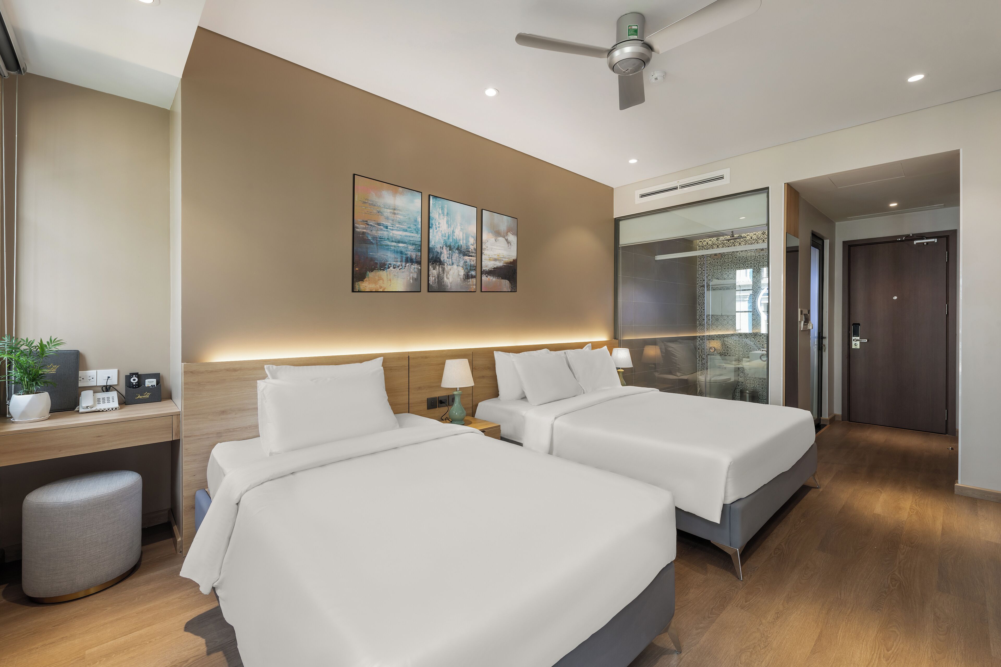 A modern hotel room with two beds, a desk, a small stool, and a ceiling fan.