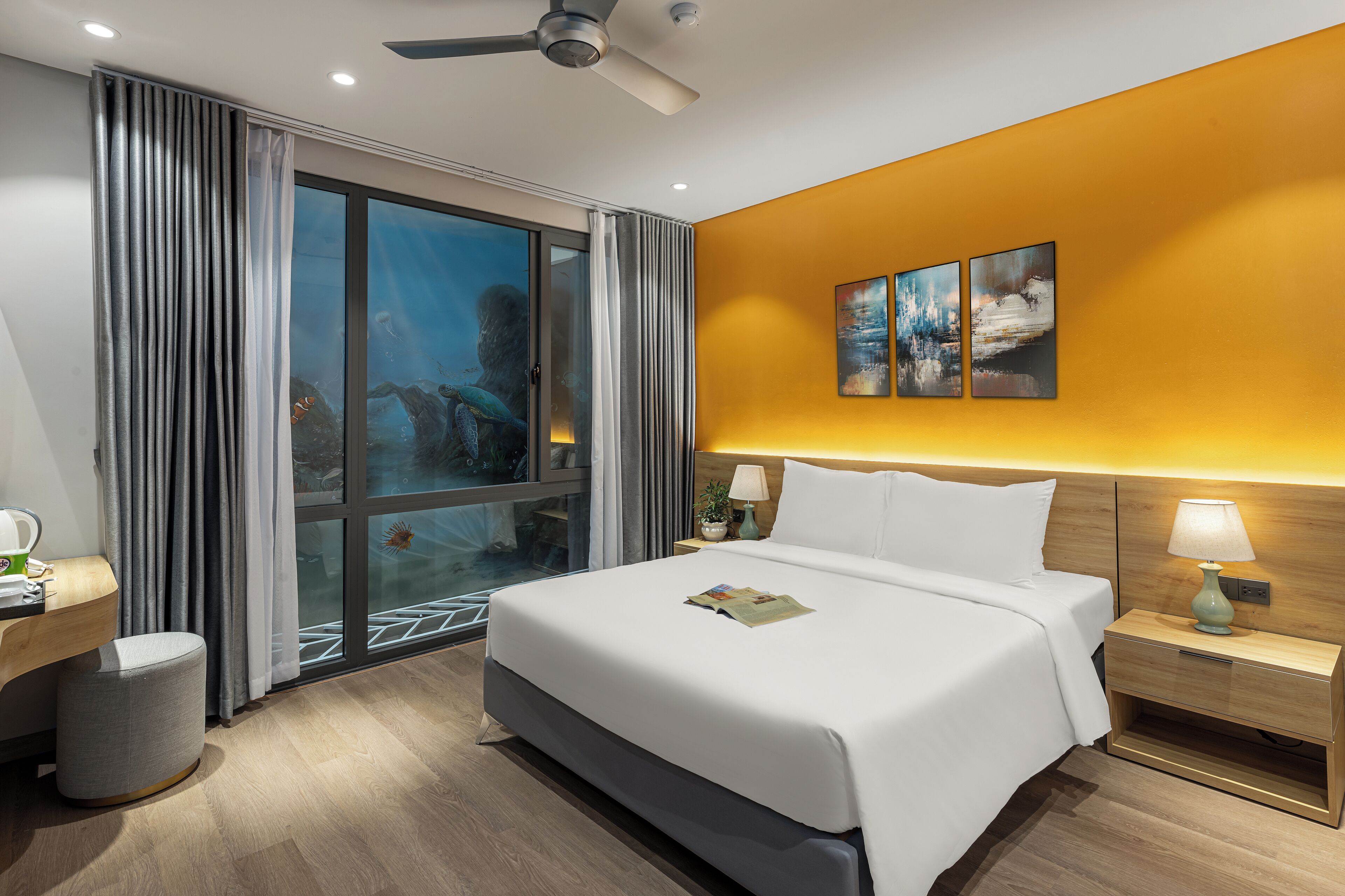Habitación de hotel con una cama grande, un escritorio, una silla, un cubo de basura, una mesita de noche con una lámpara y una ventana con cortinas.