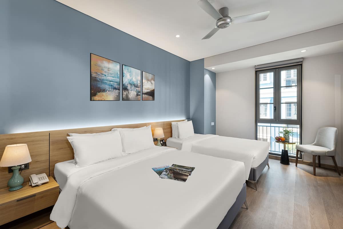 A hotel room with two beds, a ceiling fan, a window with a view, and artwork on the wall.