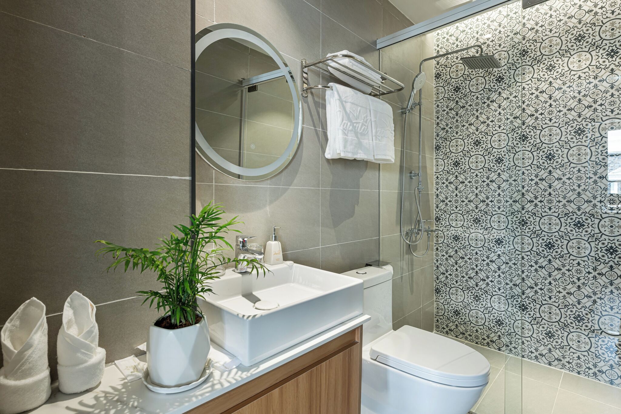 A modern bathroom with a white sink, a round mirror, a potted plant, a toilet, and a shower area with patterned tiles.