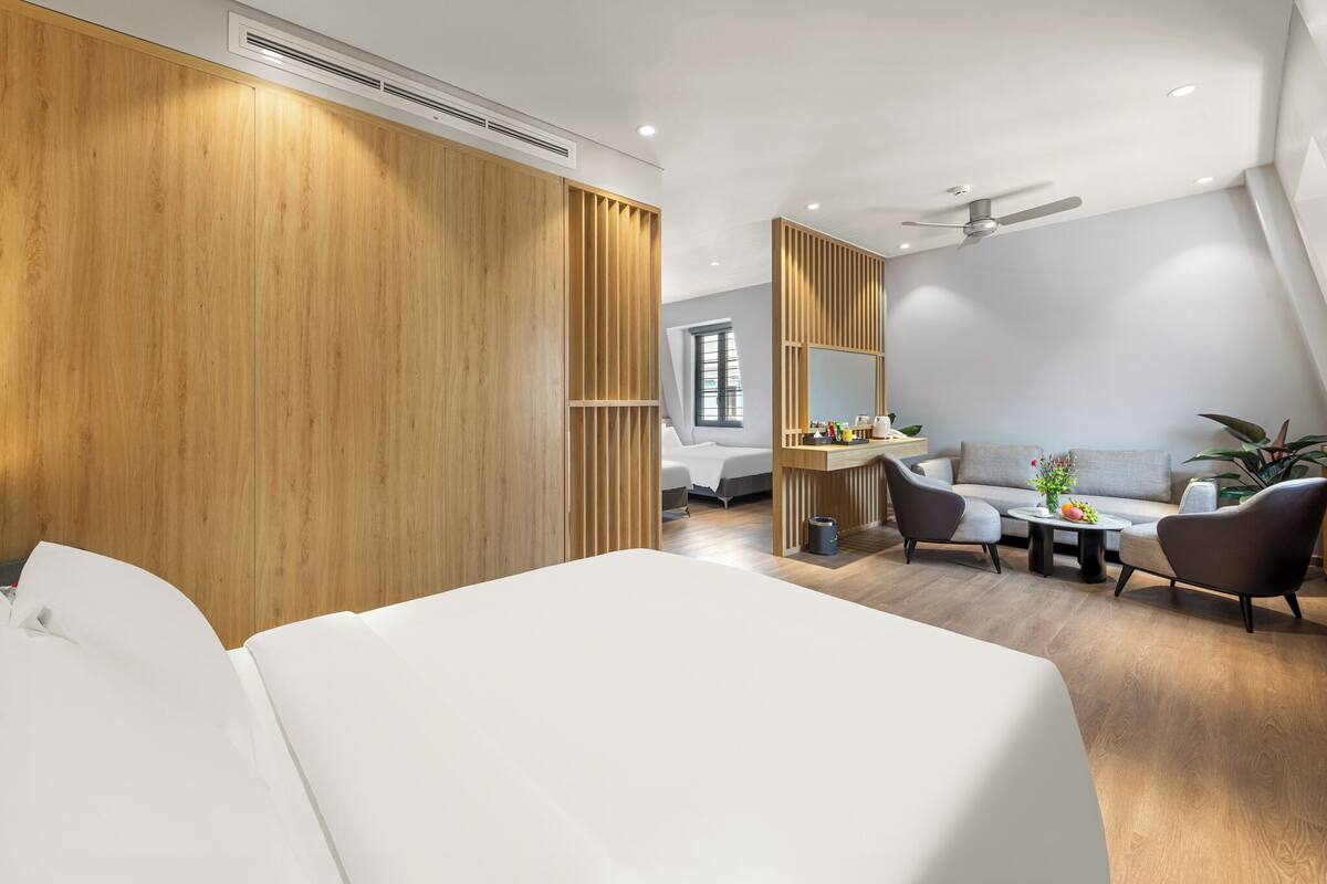 A modern hotel room with a large bed, a seating area with a sofa and chairs, a small table with a vase of flowers, and a large window providing natural light.