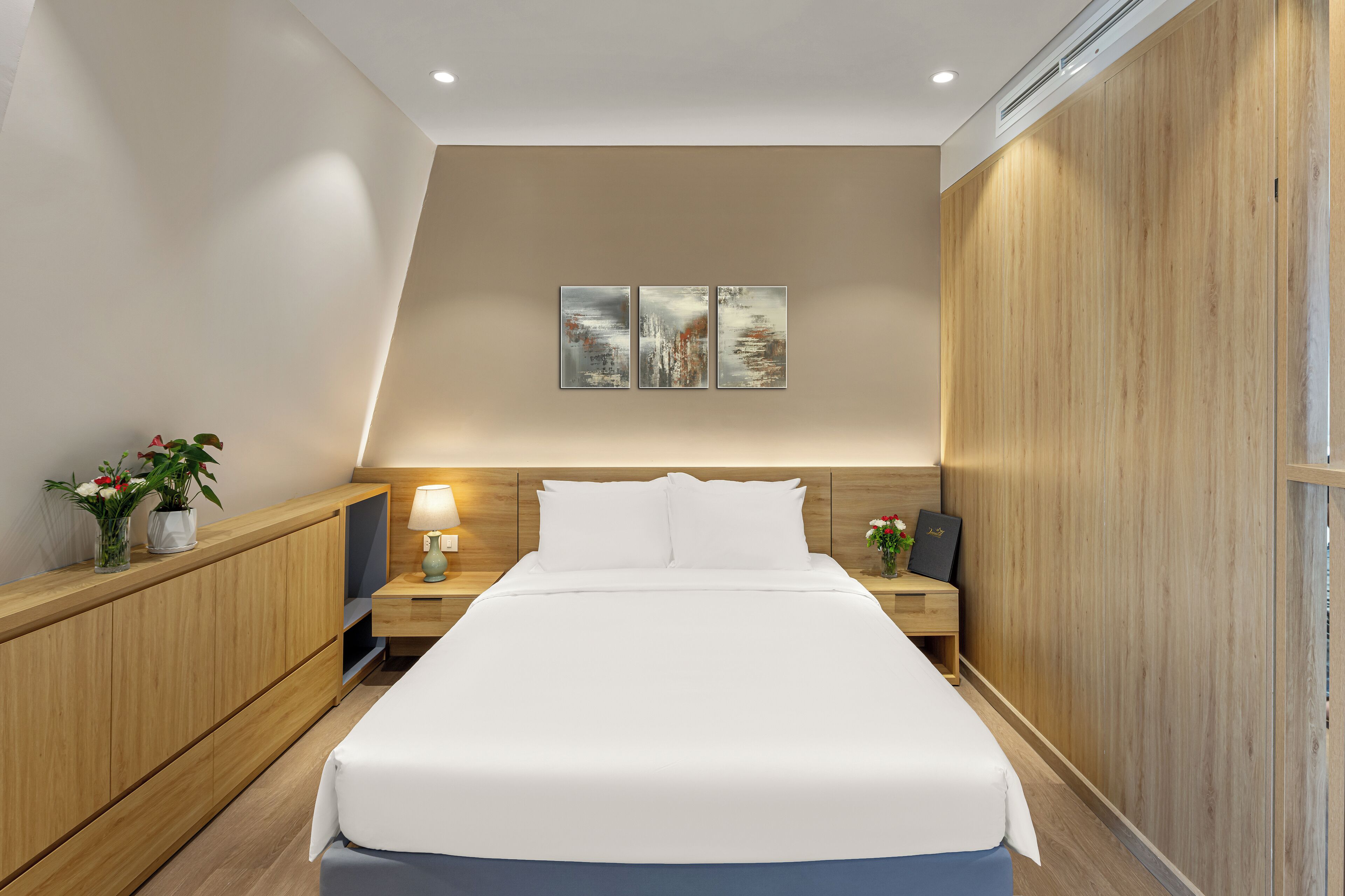 A modern hotel room with a large bed, wooden headboard, bedside tables with lamps, and a wall-mounted artwork.