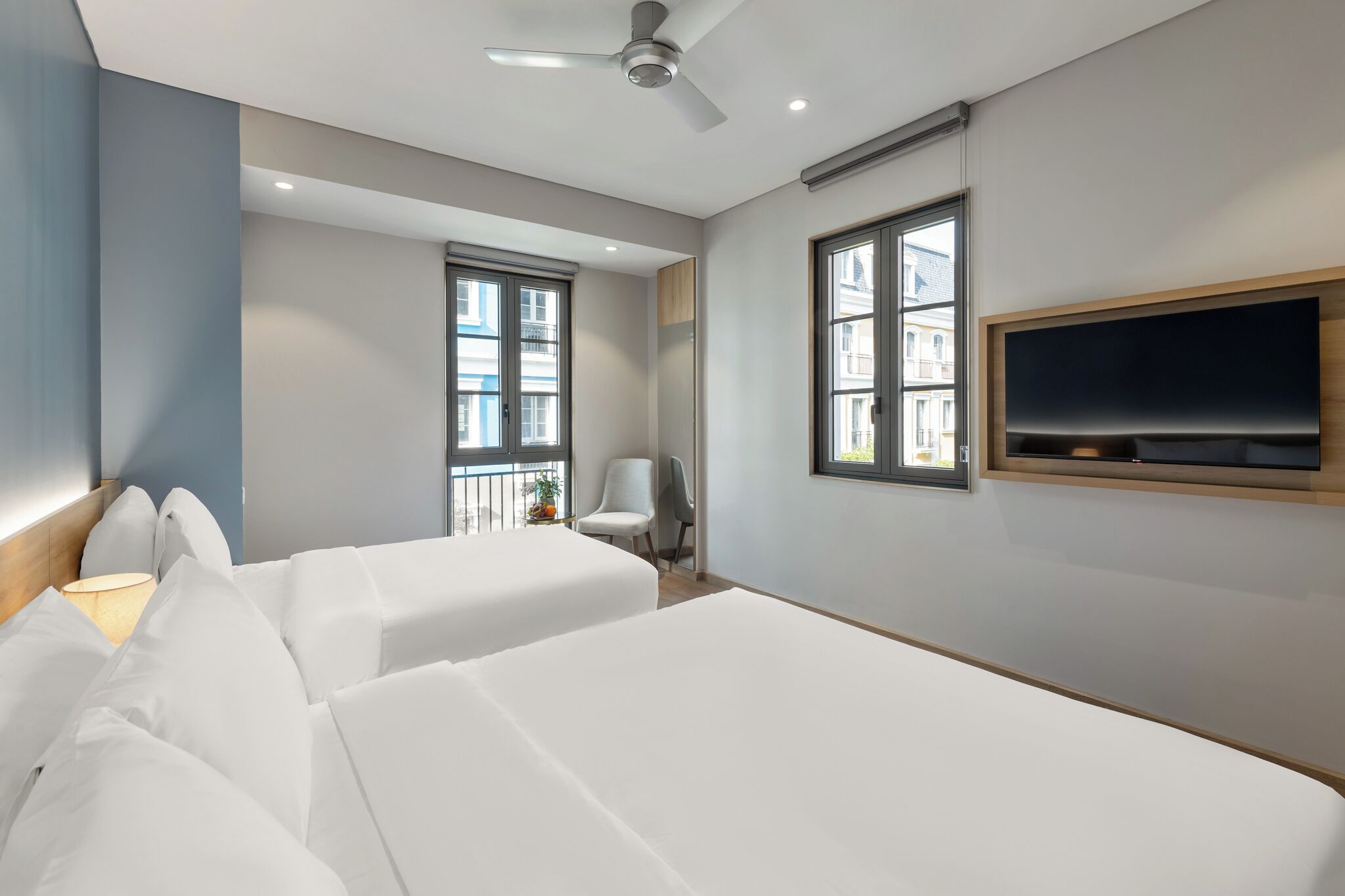 Ein Hotelzimmer mit zwei Betten, einem Deckenventilator, einem an der Wand befestigten Fernseher und großen Fenstern.