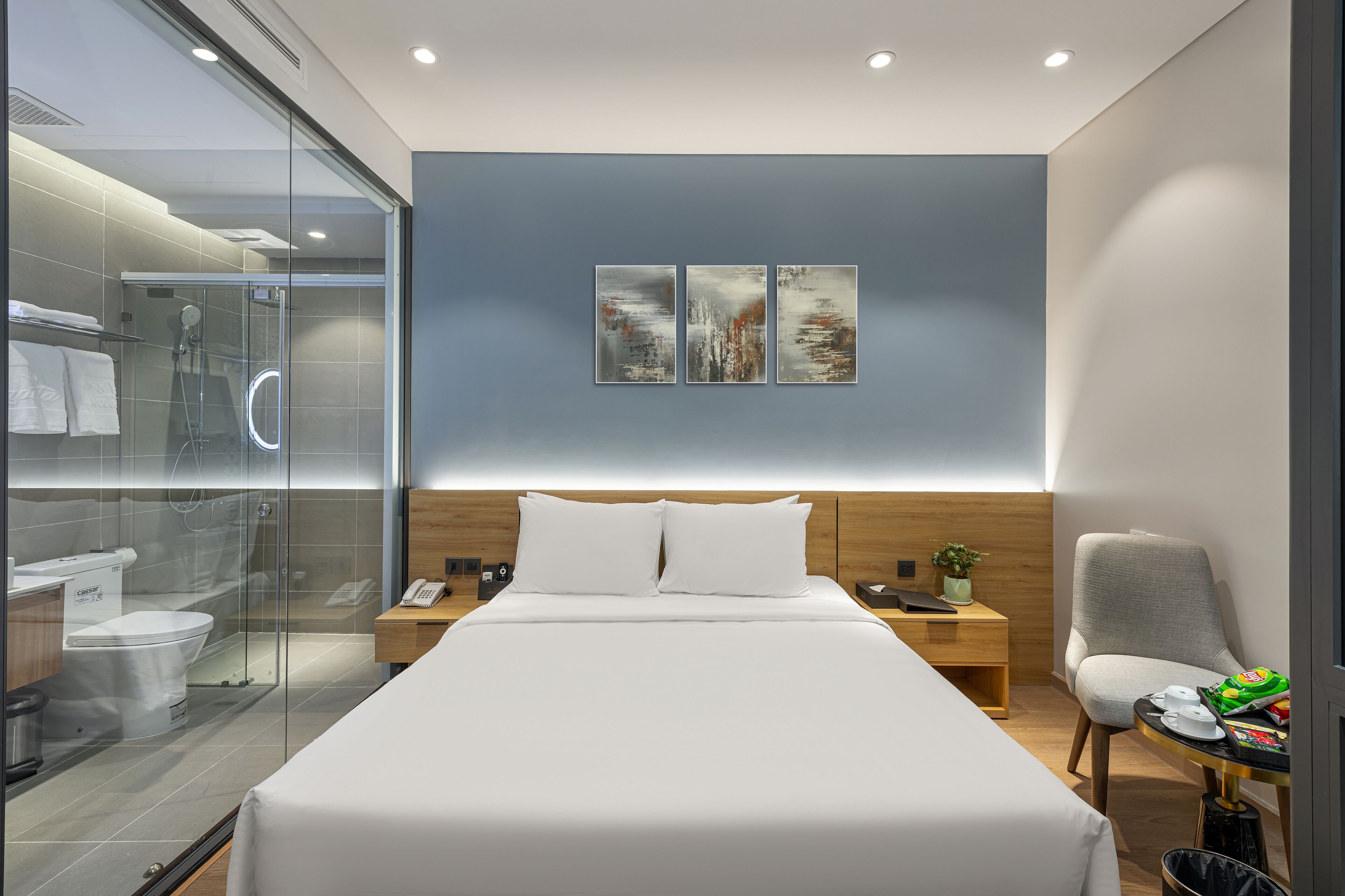 A modern hotel room with a large bed, a bedside table with a lamp, a chair, and a glass shower enclosure.