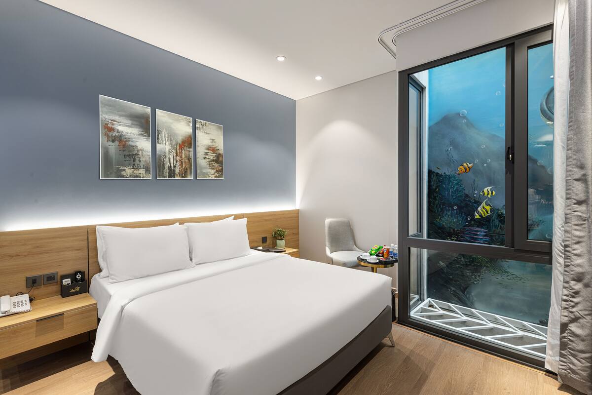 A modern hotel room with a large bed, a nightstand with a phone, a chair, and a window with an underwater view.