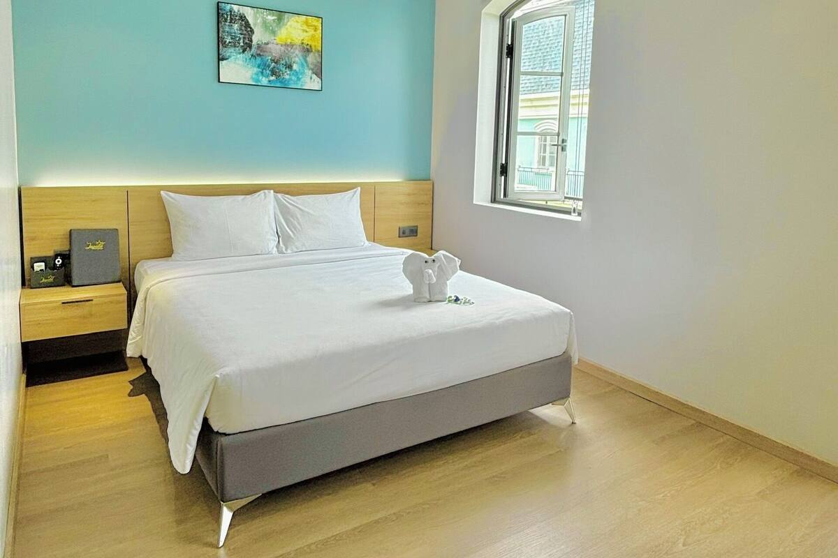A modern hotel room with a bed, a nightstand, a ceiling fan, a window, and a painting on the wall.
