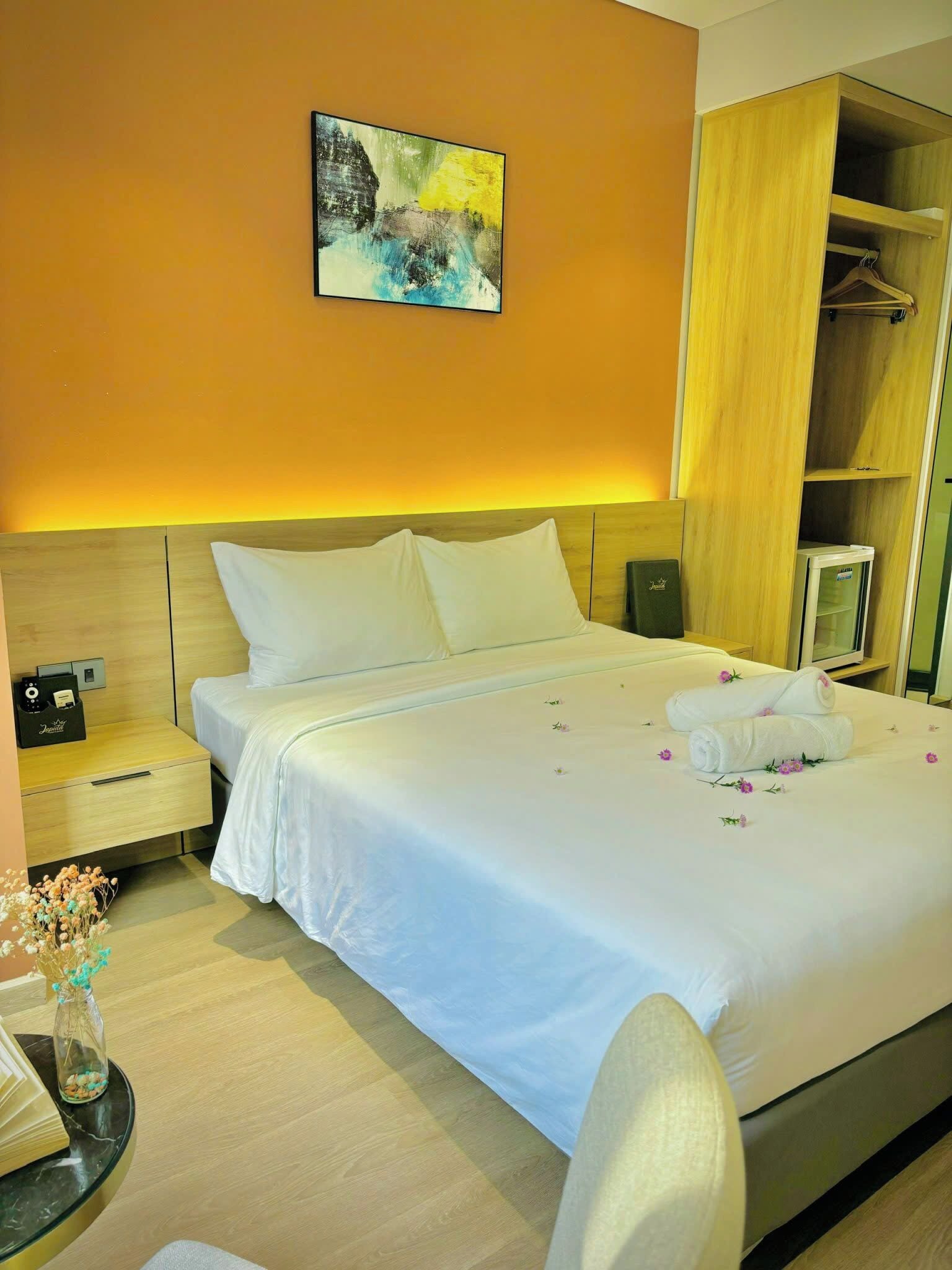 Ein Hotelzimmer mit einem ordentlich bezogenen Bett, einem Nachttisch mit Lampe, einem kleinen Tisch mit Blumenvase und einem Kleiderschrank.