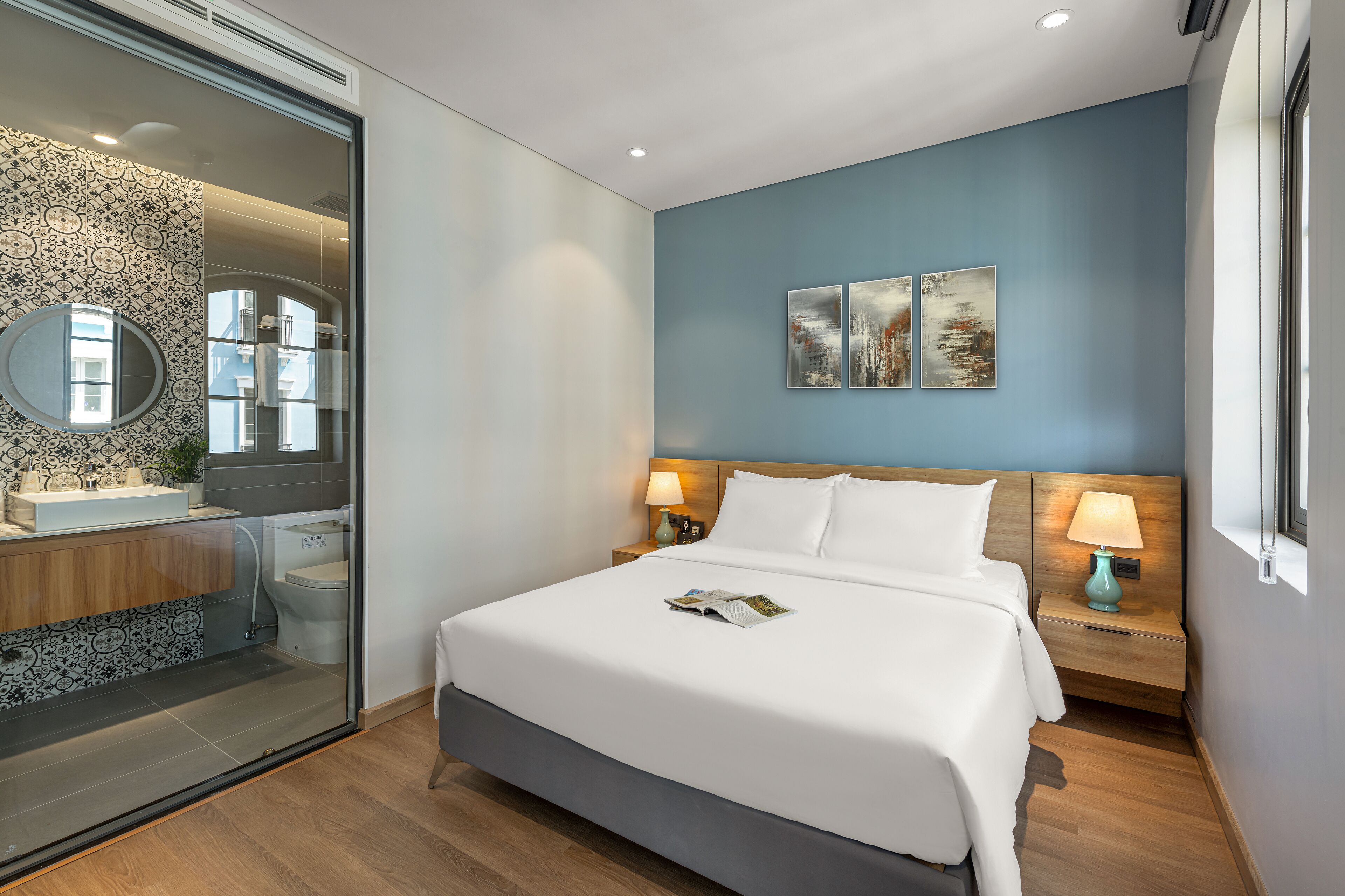 A modern hotel room with a large bed, a glass shower enclosure, a bedside table with a lamp, and artwork on the wall.