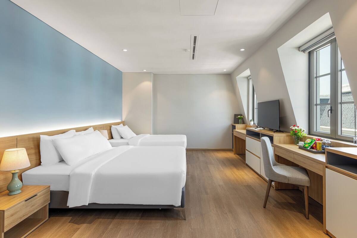 A modern hotel room with a large bed, a desk, and a chair. There is a TV on the desk and a window with a view of the city.