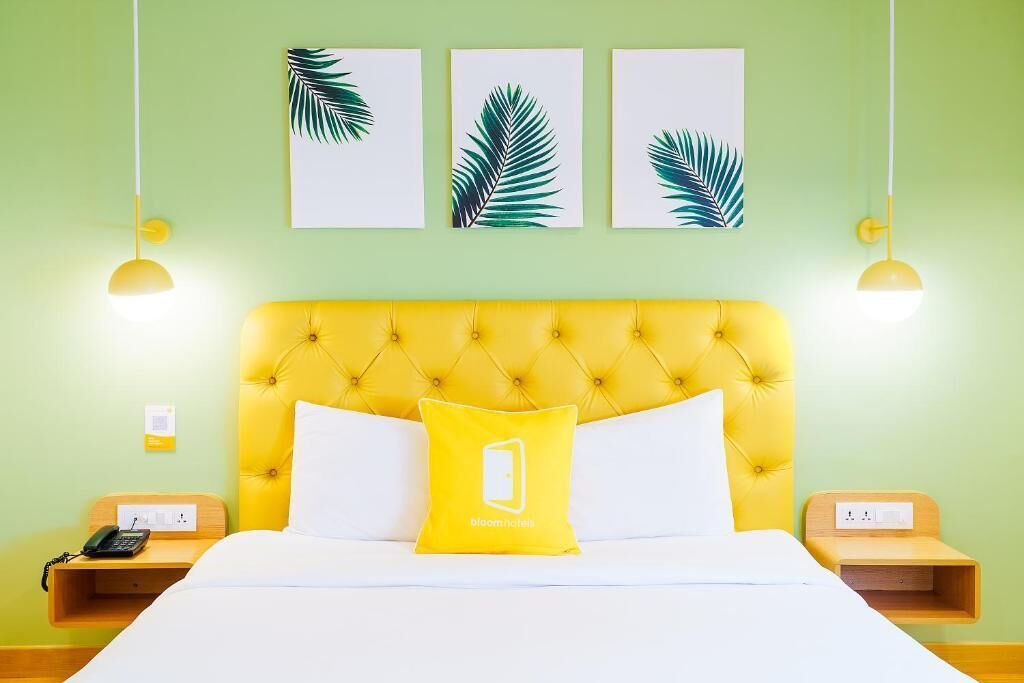 A hotel room with a yellow tufted headboard, white bedding, a yellow pillow with a logo, bedside tables with lamps, and three framed leaf artworks on the wall.