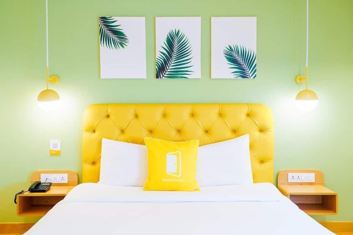 Habitación de hotel con un cabecero amarillo capitonado, ropa de cama blanca, un almohadón amarillo con un logotipo, mesitas de noche con lámparas y tres obras de arte con hojas enmarcadas en la pared.