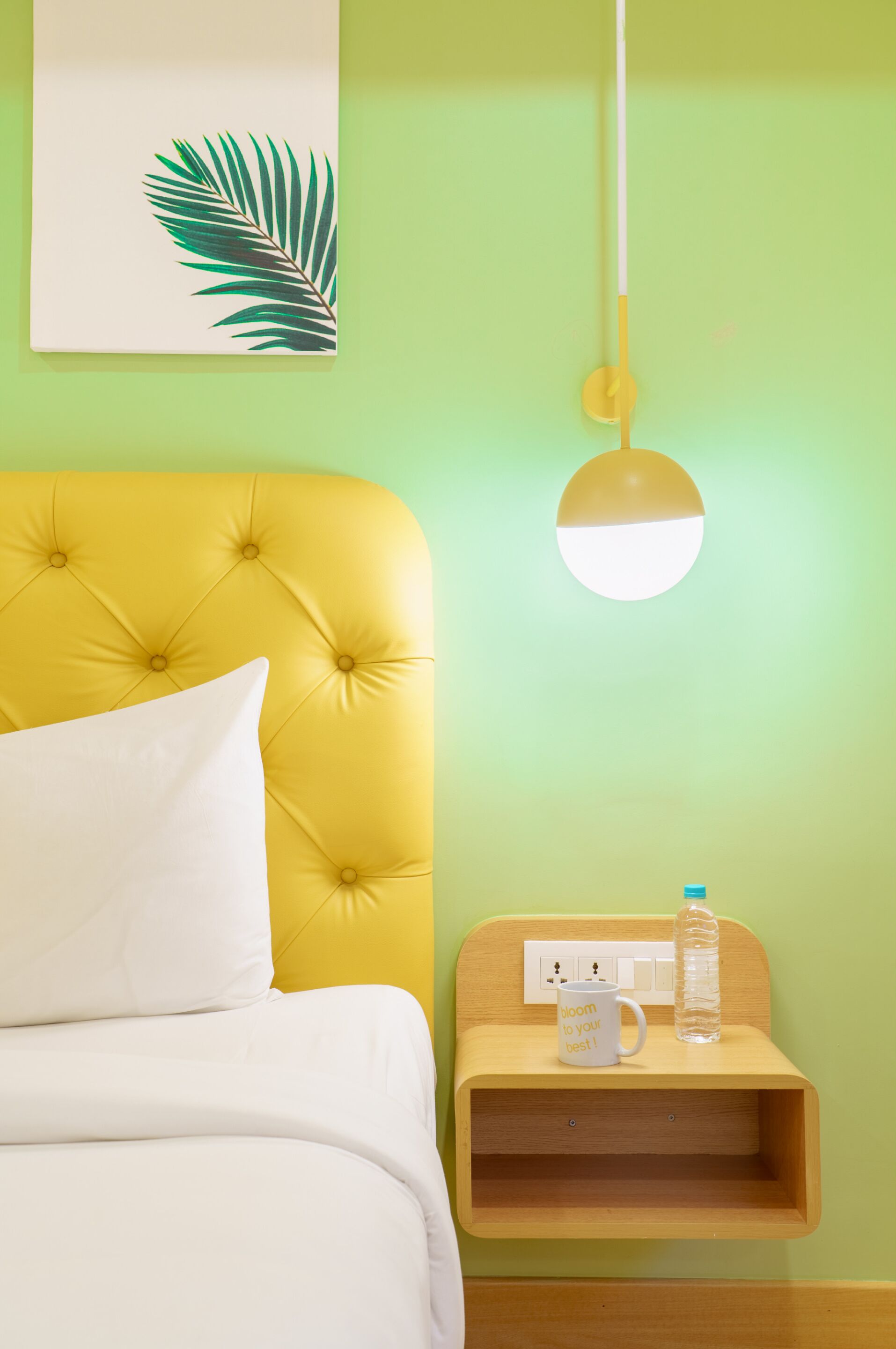 A room with a yellow tufted headboard, white bedding, a wall-mounted light fixture, and a bedside table with a mug and water bottle.