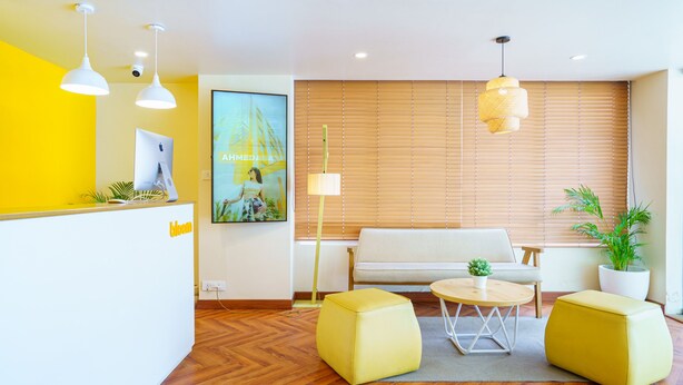 A reception area with a white reception desk, yellow ottomans, a white sofa, and a wooden floor. - Bloom Hotel - Cyber Towers (Hyderabad)