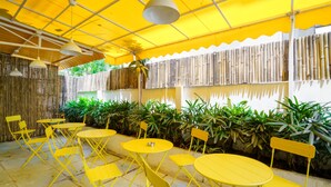 An outdoor seating area with yellow tables and chairs, a bamboo fence, and hanging pendant lights. - Bloom Hotel - Cyber Towers (Hyderabad)
