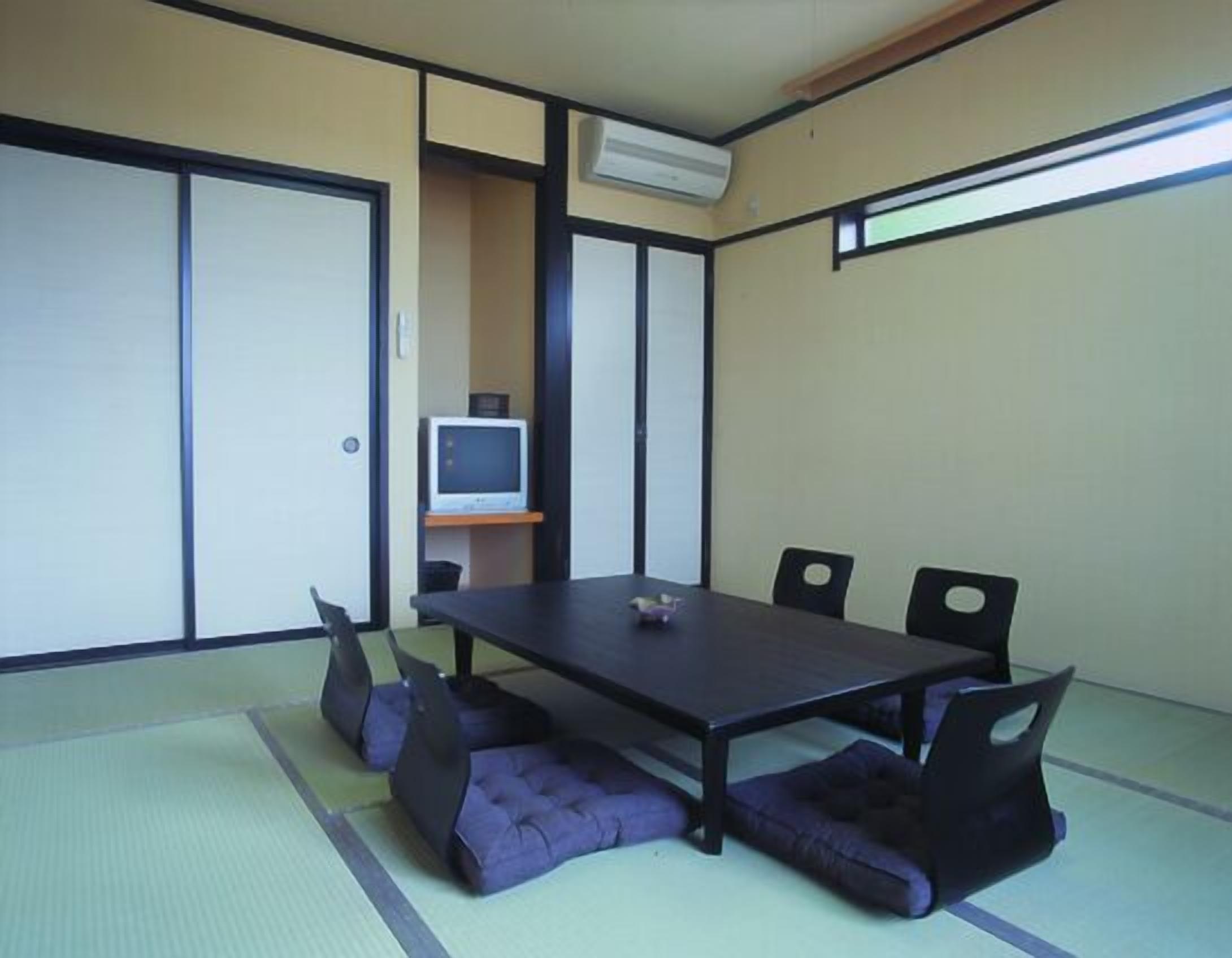 A traditional Japanese room with tatami flooring, a low table, four chairs, a television on a shelf, and sliding doors.