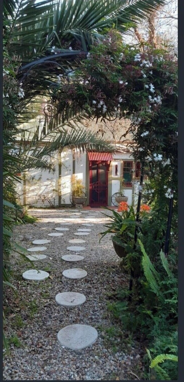 Un chemin menant à un bâtiment doté d’une porte rouge, entouré d’une végétation luxuriante et de plantes en fleurs.