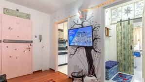A hotel lobby with a large TV screen displaying a cityscape, a pink door, a red rug, and a decorative tree mural. - Lodge & Art (Trieste)