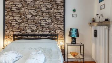 Une chambre avec une tête de lit en pierre, un lit avec du linge de lit et des serviettes blancs, une table de chevet avec une lampe et un radiateur.