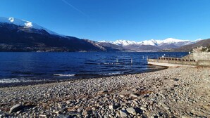 A rocky shoreline with a pier extending into a calm lake, surrounded by snow-capped mountains under a clear blue sky.