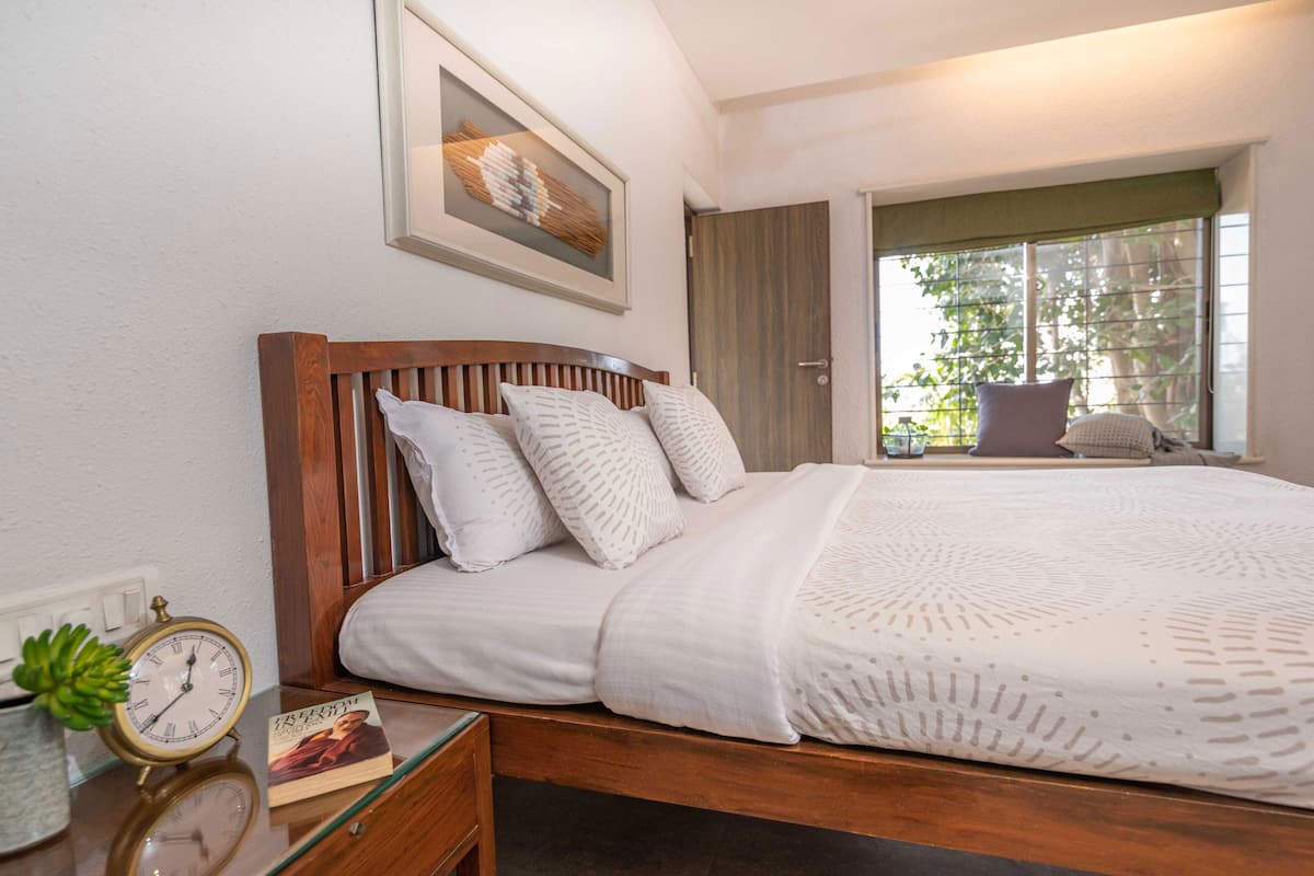 A neatly made bed with white linens and a wooden headboard, a bedside table with a clock and a book, and a window with a view of greenery outside.