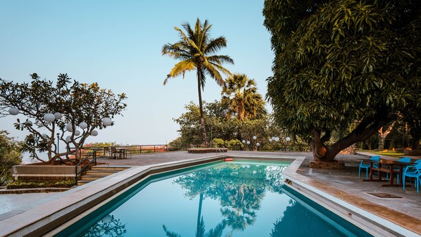 Una piscina con agua cristalina, rodeada de palmeras y un patio con sillas y mesas.