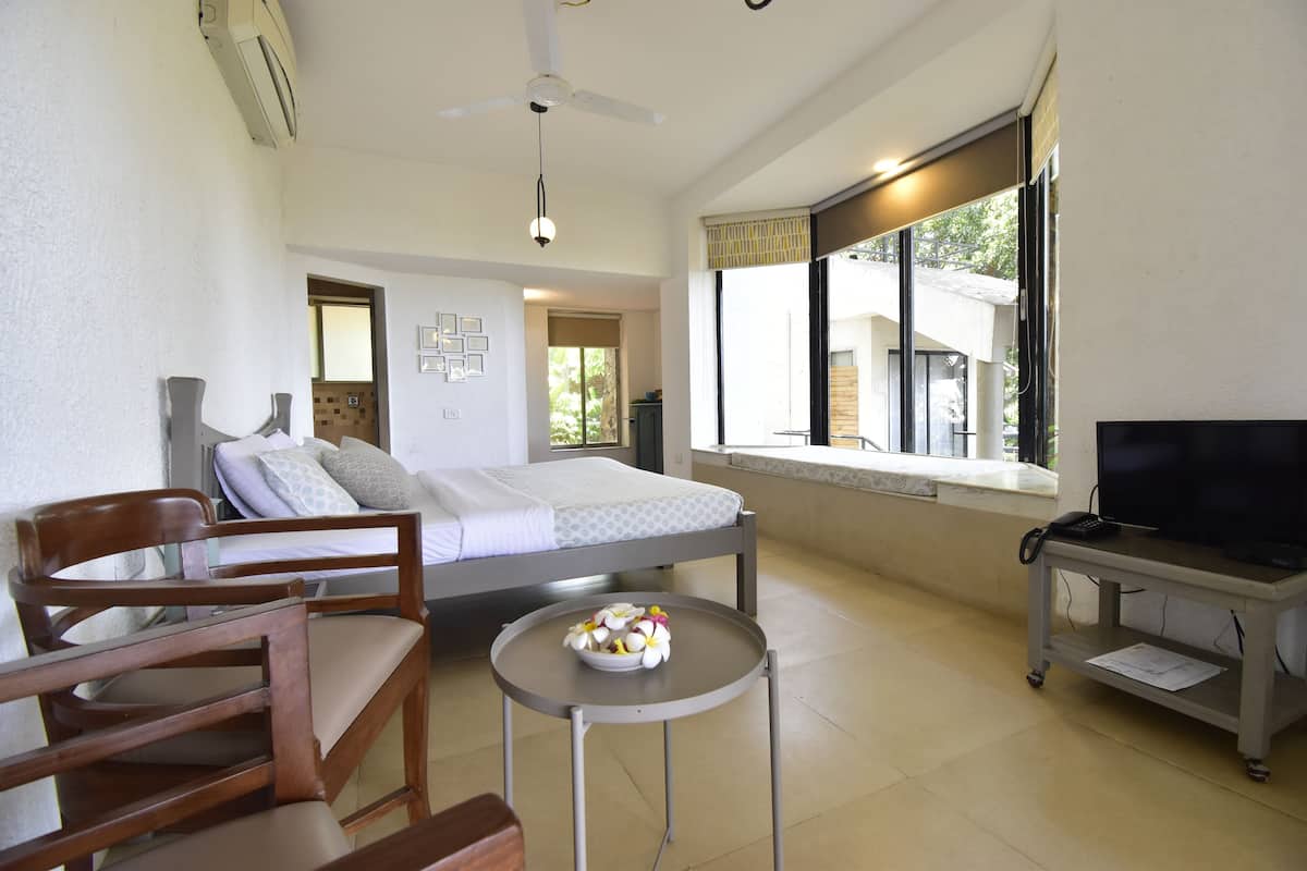 A bedroom with a bed, a TV on a stand, a desk with a chair, and a window with a view of the outside.