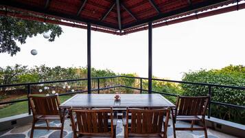 A covered outdoor seating area with a table and chairs, surrounded by stone flooring and a railing, overlooking a garden.