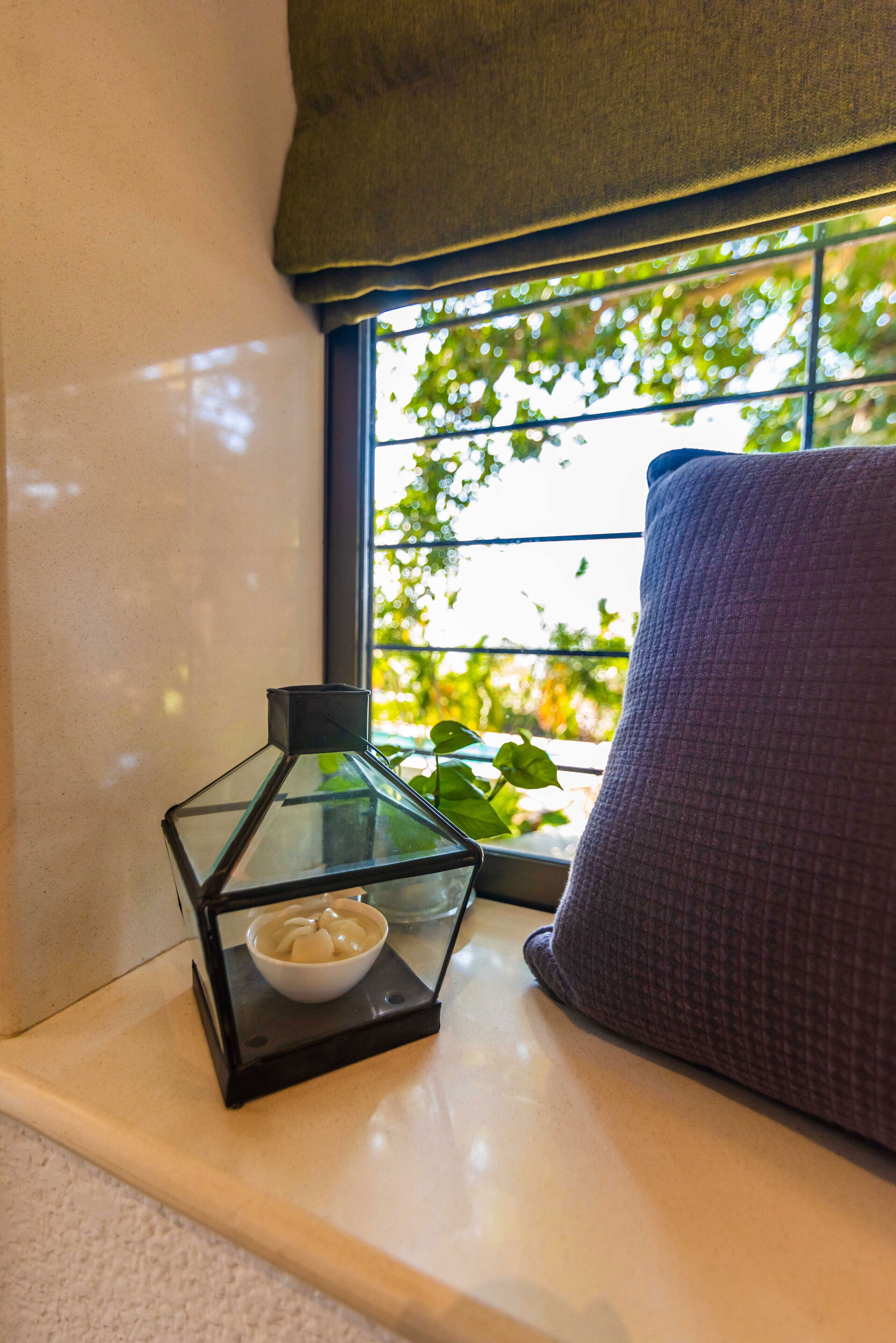 A corner of a room with a window, a cushion, and a glass lantern with a white object inside.