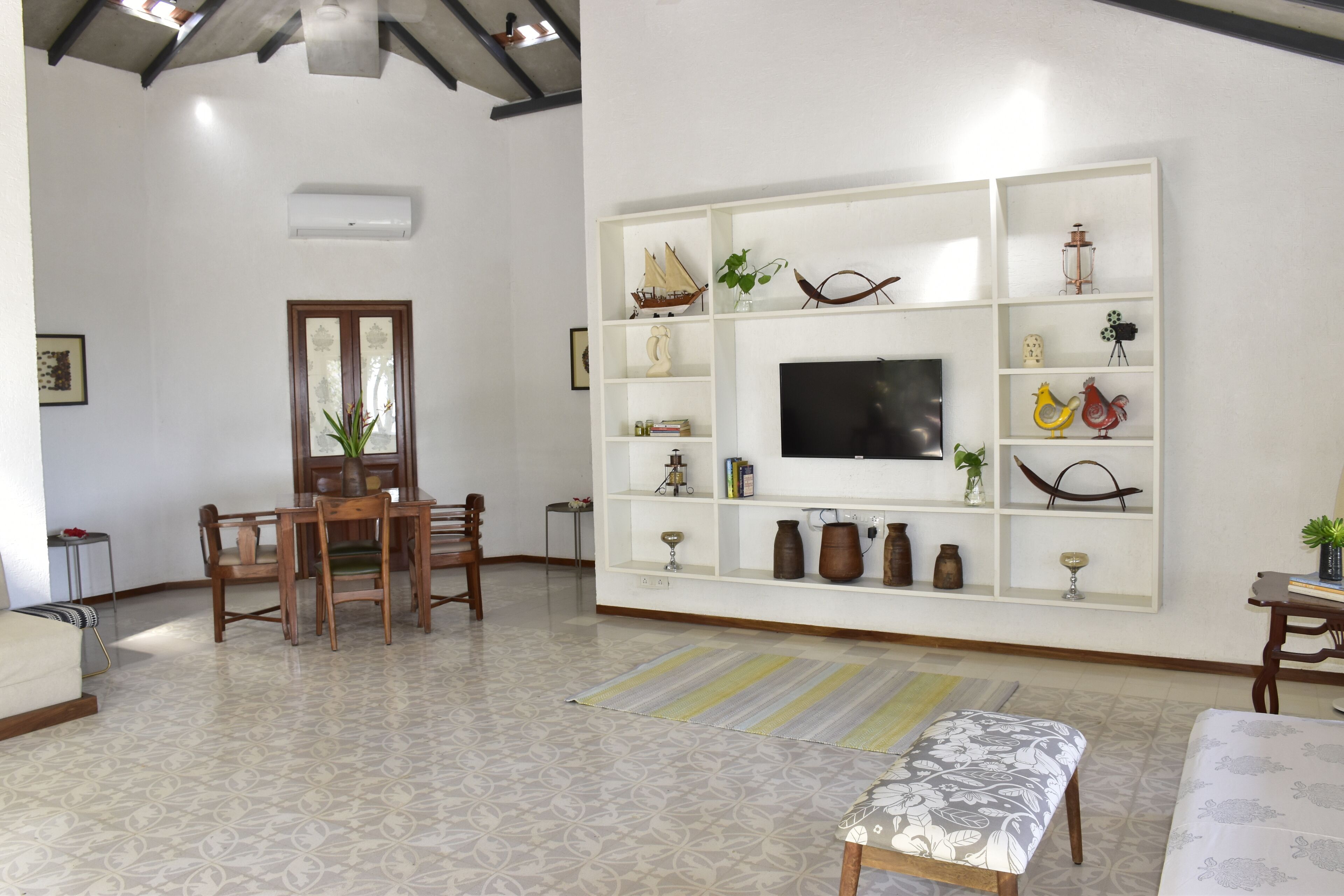 Un salon avec un téléviseur posé sur une étagère blanche, une table à manger avec des chaises, un tapis à motifs et un banc.