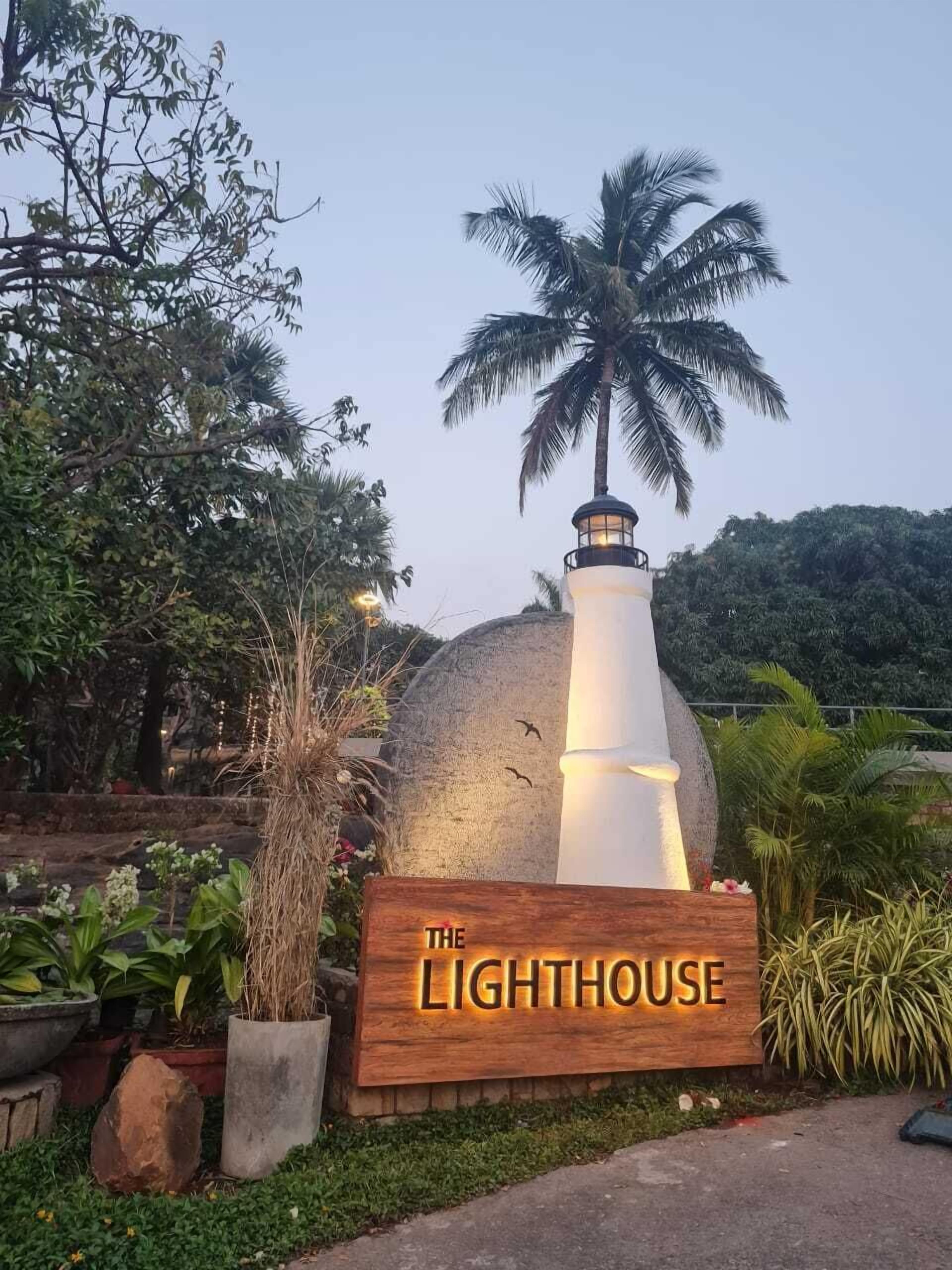 A wooden sign with "THE LIGHTHOUSE" written on it, surrounded by greenery and a palm tree.