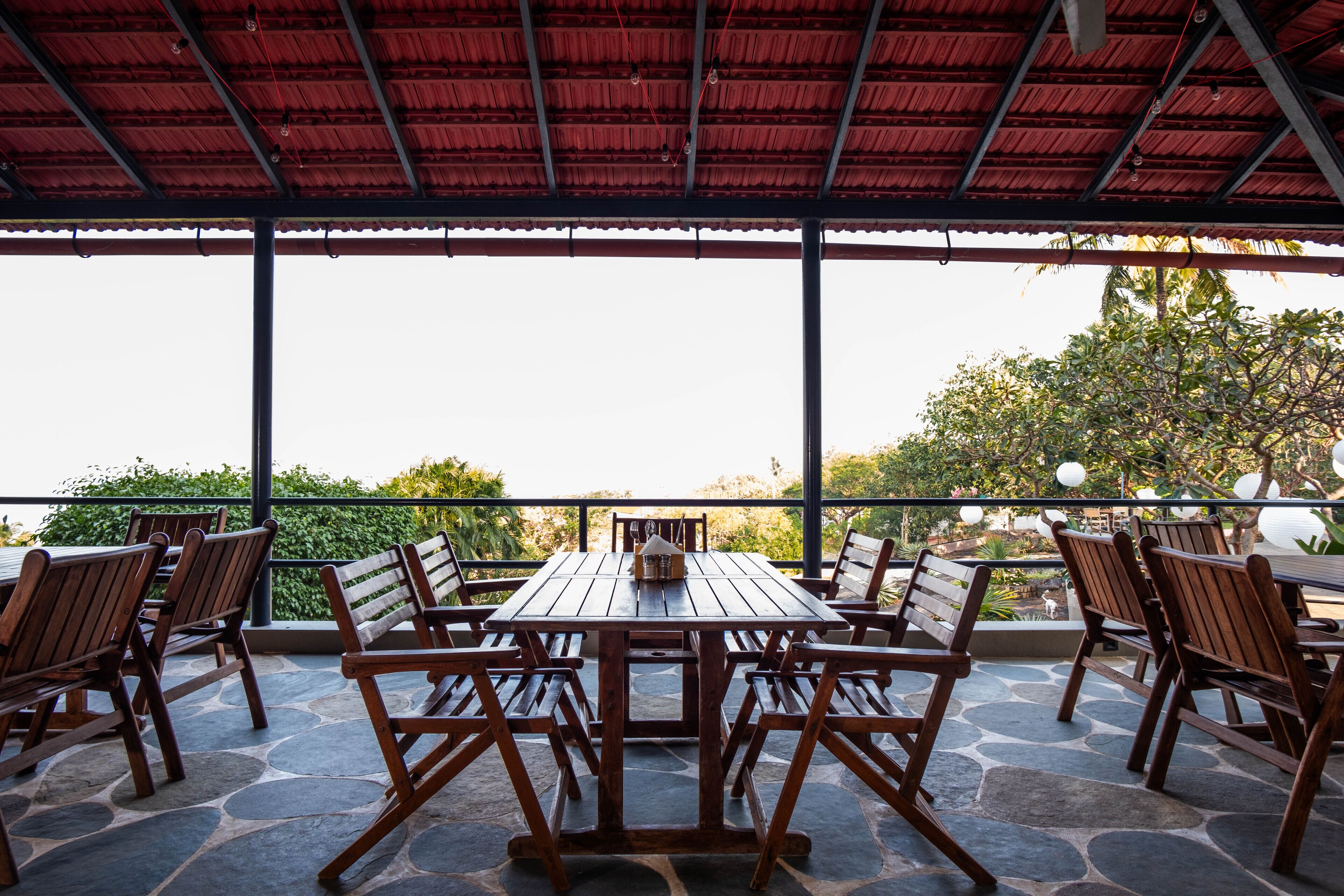 An outdoor patio with a wooden table and chairs, a red roof, and a view of greenery.