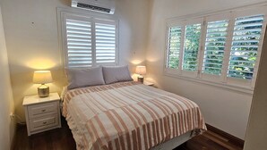 A bedroom with a bed, two bedside tables, a window with blinds, and an air conditioning unit. - Magnetic Mango Escape (HORSESHOE BAY)