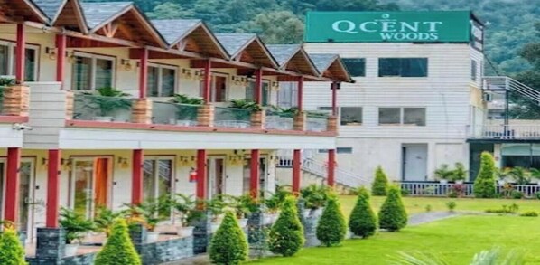 Un hotel de varios pisos con un balcón en el segundo piso, un letrero que dice 'QCent Woods', y un césped bien cuidado con arbustos recortados.