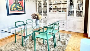 Ein Essbereich mit einem Glastisch, blaugrünen Stühlen, einer Kristallleuchter, einem Schrank mit Glastüren und einem gerahmten Gemälde.