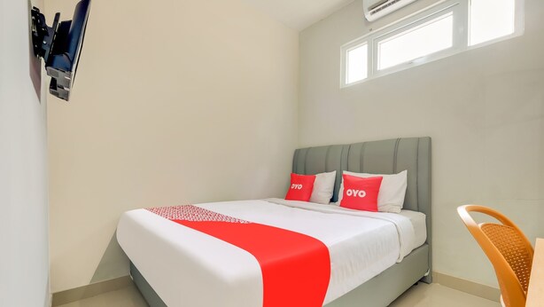 Una cama bien hecha con una colcha roja y blanca, dos almohadas con la marca 'OYO', un cabecero tapizado gris y una mesita de noche con una silla en la esquina.