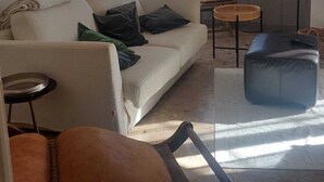 Een woonkamer met een bank, fauteuil, voetenbankje en een klein tafeltje. Er hangen afbeeldingen in lijsten aan de muur en een raam met gordijnen.