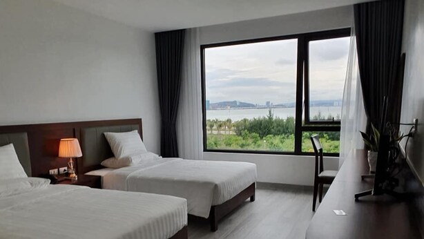 Una camera d'albergo con due letti, una scrivania e una grande finestra che si affaccia su spazi verdi e un panorama urbano in lontananza.