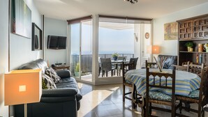 A room with a sofa, a dining table with chairs, a TV mounted on the wall, and a view through a sliding glass door to the outside. - Apartment Grand Costa Brava (Canyelles)