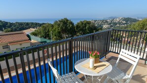 A balcony with a pool, white chairs, a table with a vase of flowers, and a view of the sea and town. - Apartment Grand Costa Brava (Canyelles)