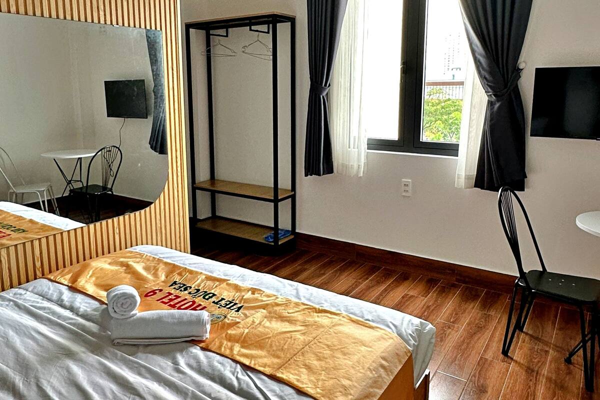 A hotel room with a bed, a wooden floor, a TV mounted on the wall, and a window with curtains.